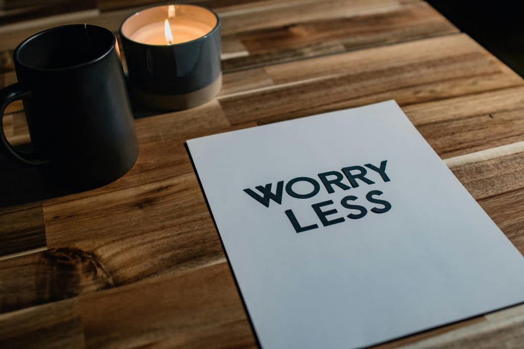 "A calming setup featuring a wooden table with a lit candle, a black mug, and a piece of paper with the phrase 'WORRY LESS' printed in bold, symbolizing mindfulness and stress relief."