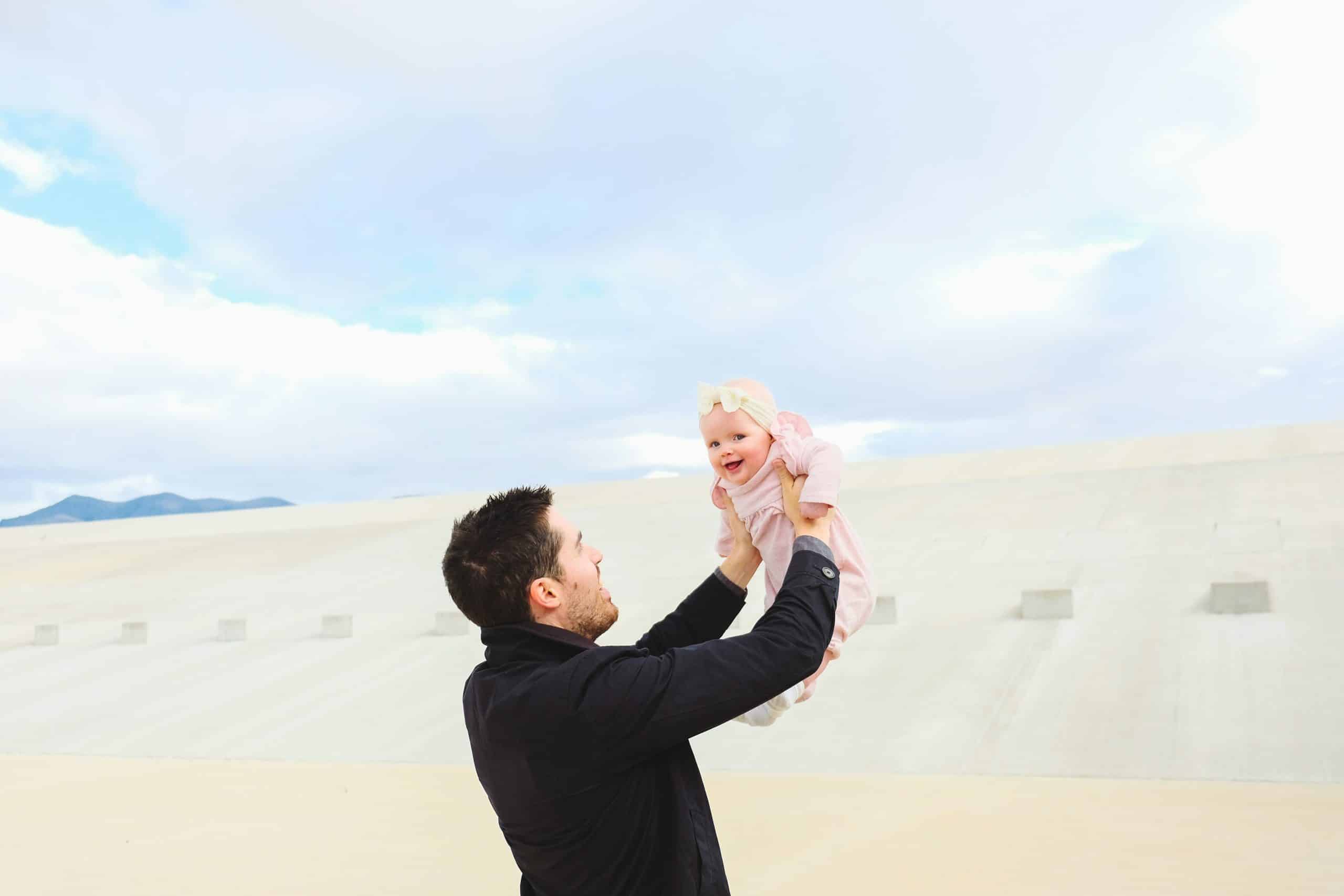 Smiling father lifts his baby daughter in the air during a bright outdoor moment, capturing joy and connection in early parenthood.